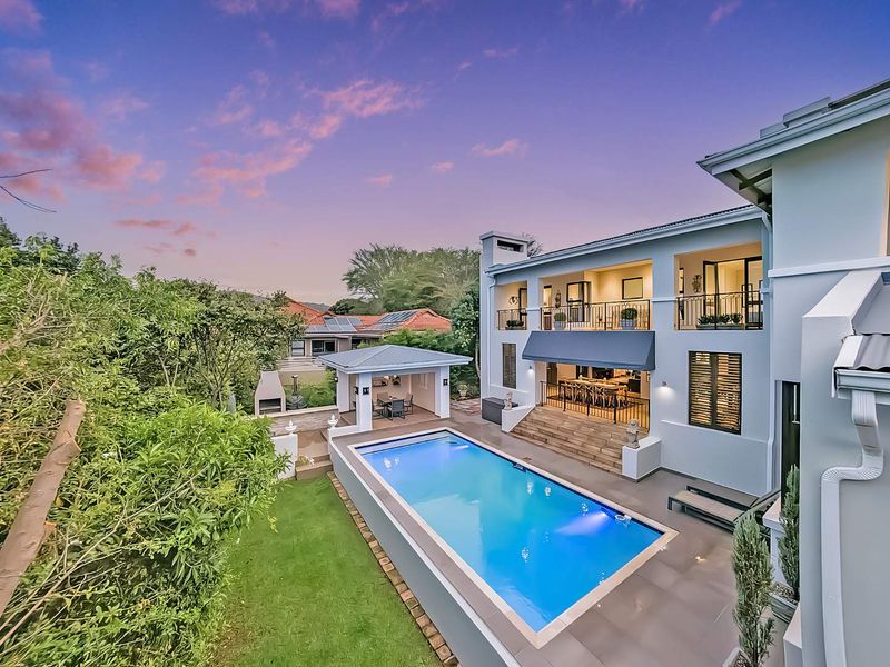 Stunning rear exterior view featuring a swimming pool, covered patio with built-in braai, and a spacious balcony overlooking the landscaped garden.