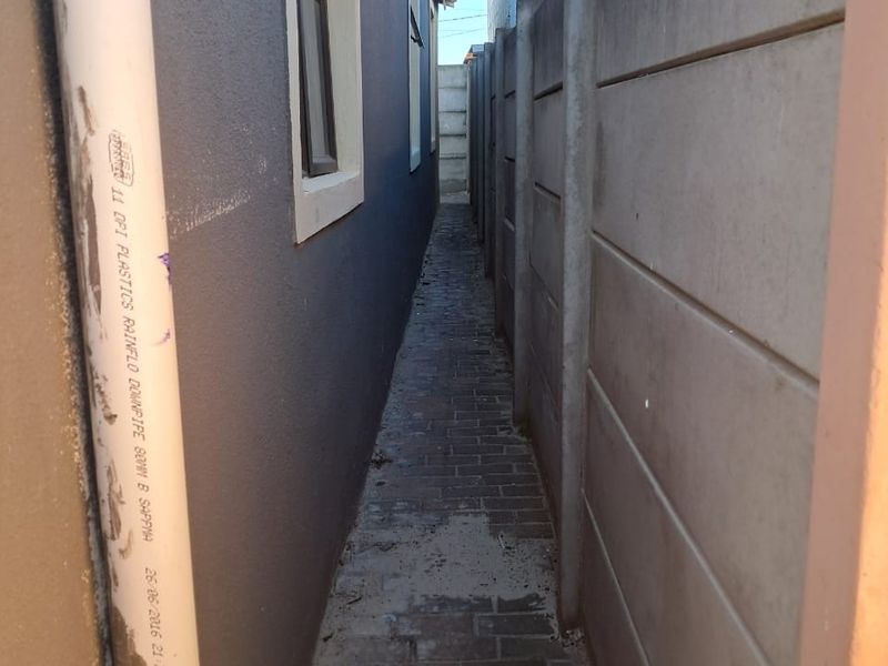 A narrow side passage featuring a paved walkway, precast concrete boundary wall, and exterior wall with a rainwater downpipe.