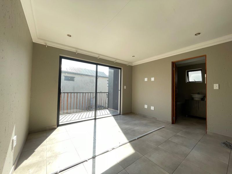 A bright, empty bedroom featuring tiled floors, recessed lighting, and a sliding glass door leading to a private balcony, with an en-suite bathroom attached.