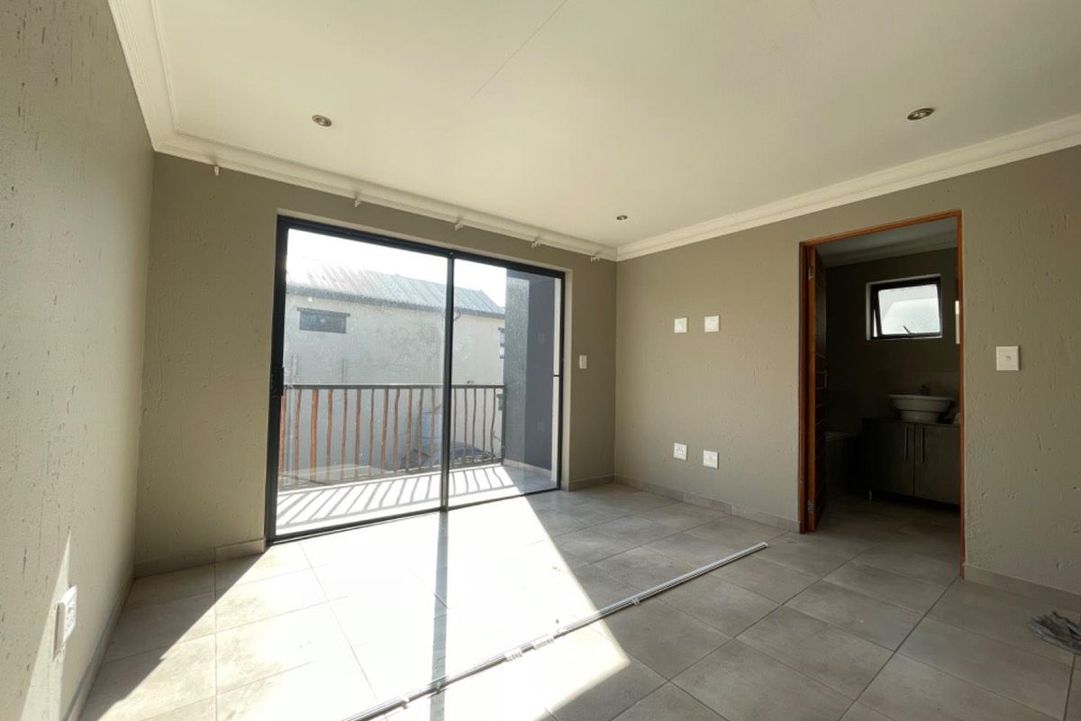 A bright, empty bedroom featuring tiled floors, recessed lighting, and a sliding glass door leading to a private balcony, with an en-suite bathroom attached.
