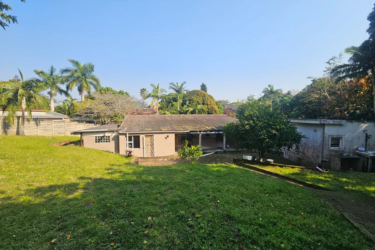 Rear view of the property showing a single-story house with a tiled roof, concrete patio area, and a large surrounding garden with boundary walls.
