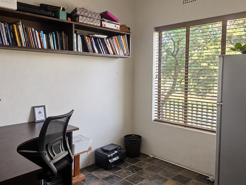 A functional home office space featuring built-in wall shelving, tiled flooring, and large windows with venetian blinds providing natural light.