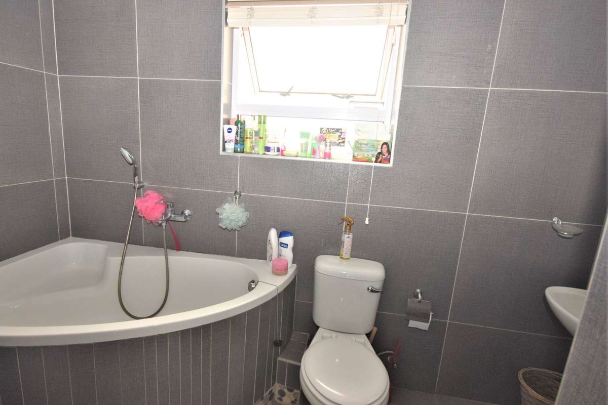 A compact bathroom featuring grey wall tiling, a corner bathtub with a handheld shower attachment, a toilet, and a wall-mounted basin.