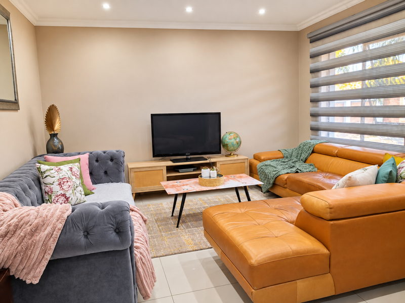 A well-maintained living room featuring neutral wall tones, tiled flooring, recessed downlighting, and modern roller blinds.