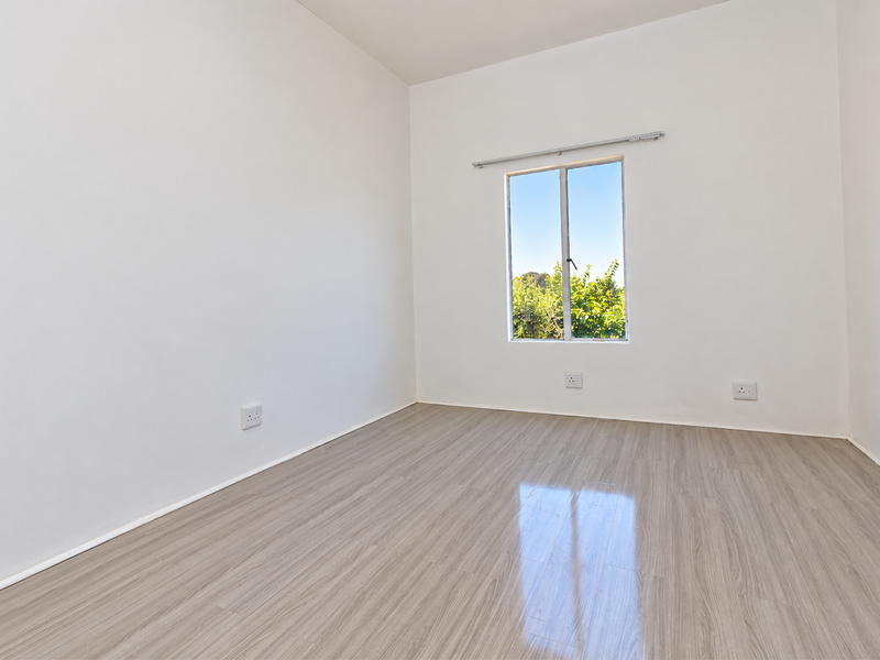 A bright, empty bedroom featuring modern laminate flooring, neutral white walls, and a window providing natural light.