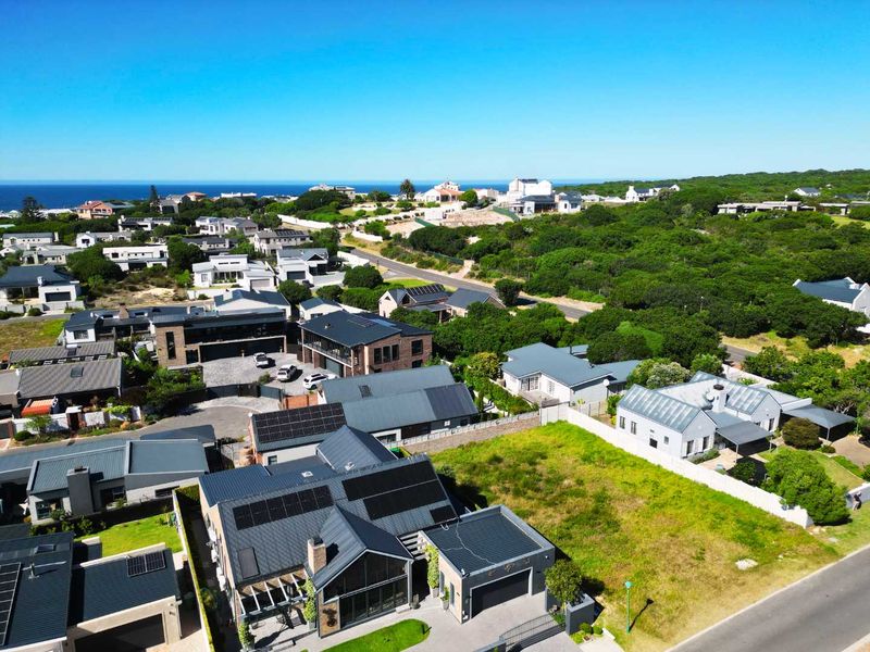 Aerial view of a residential neighborhood featuring modern homes with solar installations, paved driveways, and surrounding coastal landscape.