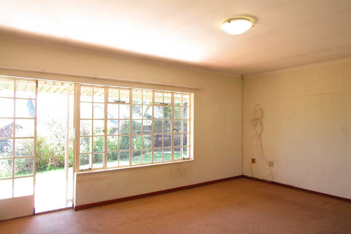 An empty, sunlit living room featuring large windows, a sliding glass door to the garden, and carpeted flooring, ready for renovation.