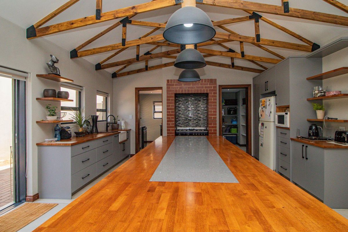 Spacious modern kitchen featuring a large central island with timber countertop, exposed timber roof trusses, and integrated cabinetry with a brick-accented cooking area.