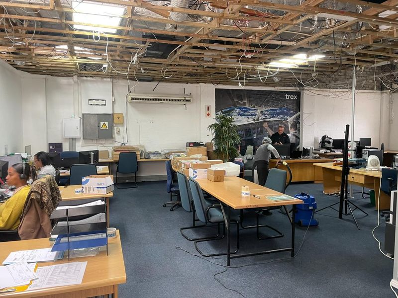 Open-plan industrial office space featuring exposed ceiling joists, wall-mounted air conditioning, and carpeted flooring, currently utilized as a functional workspace.