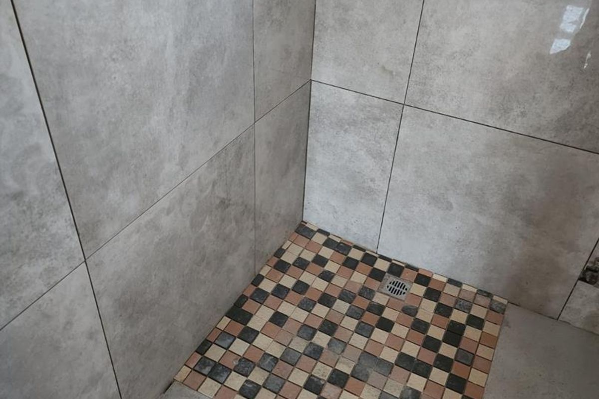 A shower enclosure featuring large format grey wall tiles and a decorative mosaic tiled floor, currently requiring finishing touches and grout maintenance.