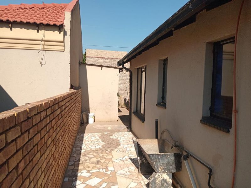 A narrow side exterior area featuring a brick boundary wall, tiled pathway, and an external utility sink attached to the main building structure.