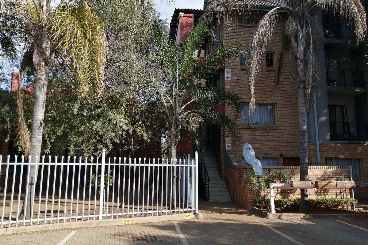 Exterior view of a multi-story brick apartment building featuring paved parking, metal security fencing, and external stair access.