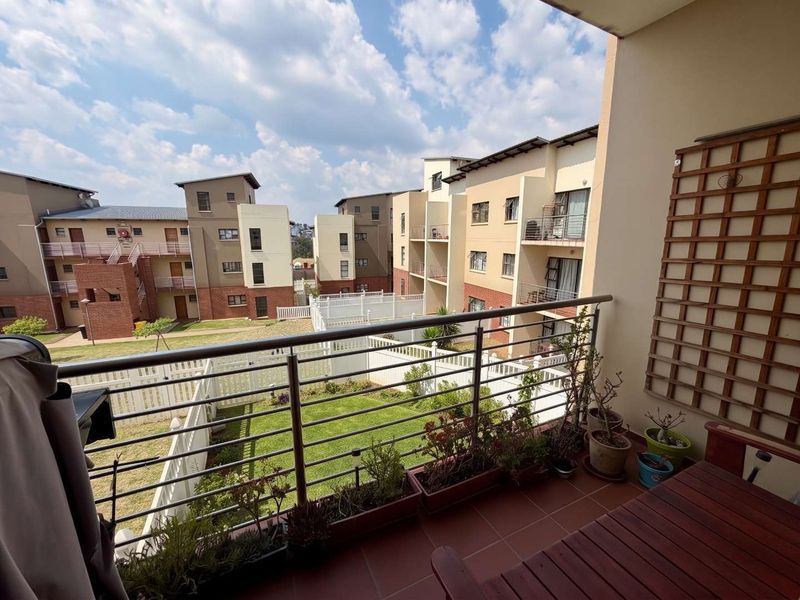 A private balcony featuring tiled flooring, a stainless steel balustrade, and a wooden wall trellis, overlooking the apartment complex grounds.
