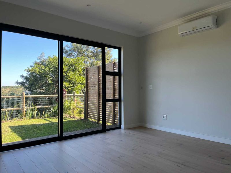 A bright, empty living room featuring modern laminate flooring, a split-system air conditioner, and large sliding glass doors opening to a private garden area.