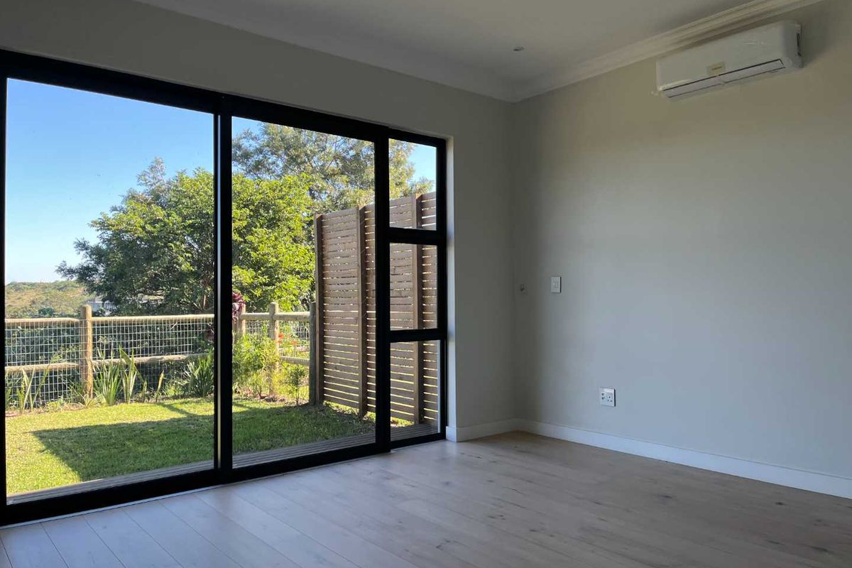 A bright, empty living room featuring modern laminate flooring, a split-system air conditioner, and large sliding glass doors opening to a private garden area.