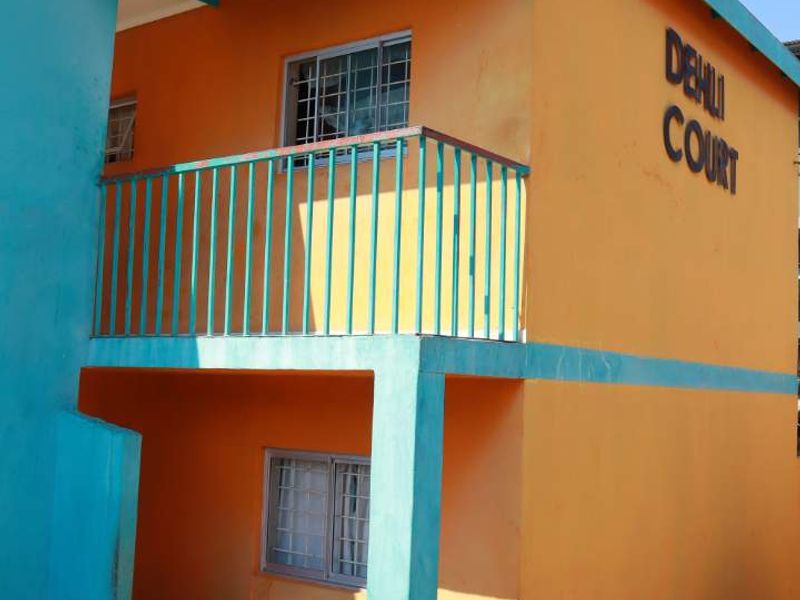 Exterior view of Dehli Court apartment building featuring orange and teal facade, metal balcony railing, and security window bars.
