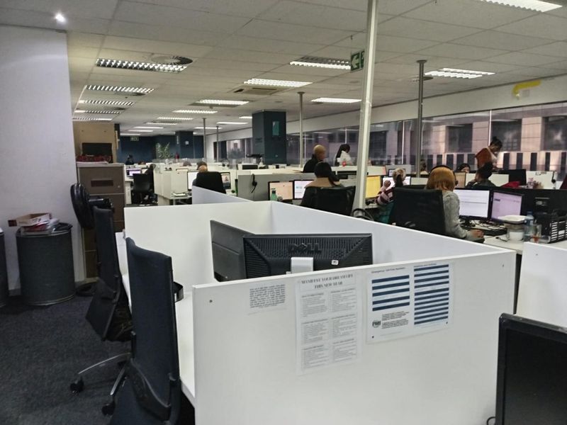 A large open-plan office space featuring suspended ceilings, recessed lighting, carpeted floors, and multiple built-in workstation partitions with large windows.