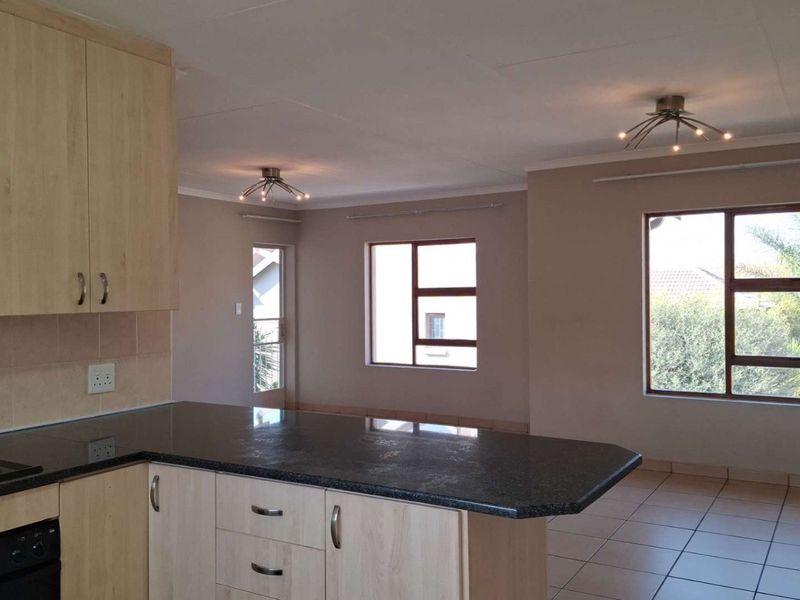 An open-plan living and kitchen area featuring light wood cabinetry, granite countertops, tiled flooring, and large windows providing natural light.