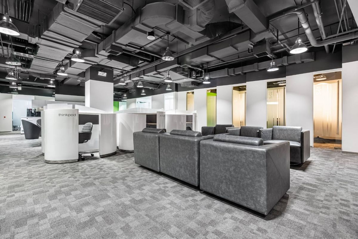 Modern open-plan office space featuring exposed industrial ceiling ductwork, carpeted flooring, and integrated private work pods with glass partition doors.