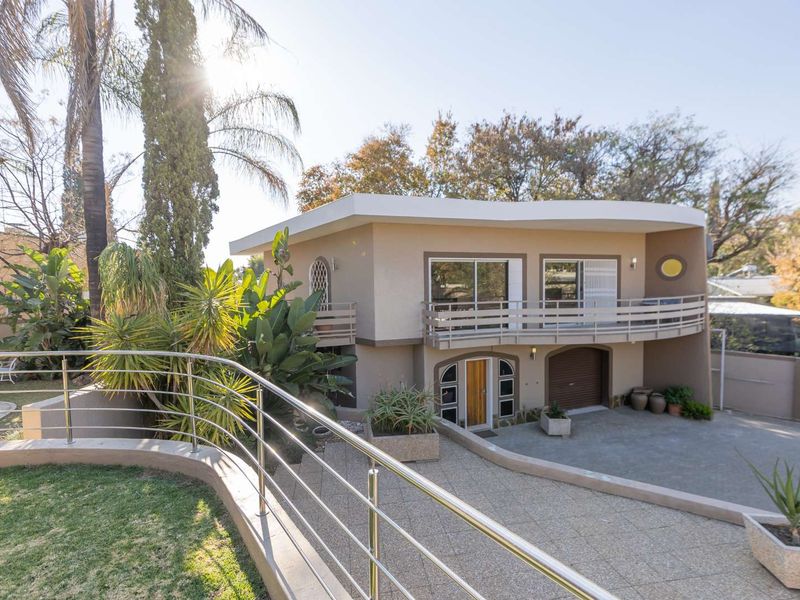 A multi-level residential property featuring a curved balcony, paved driveway, integrated garage, and a swimming pool set within a landscaped garden.