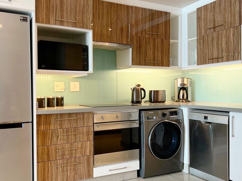Modern kitchen featuring wood-grain cabinetry, glass splashback, built-in oven, hob, extractor fan, and integrated space for laundry and dishwasher appliances.