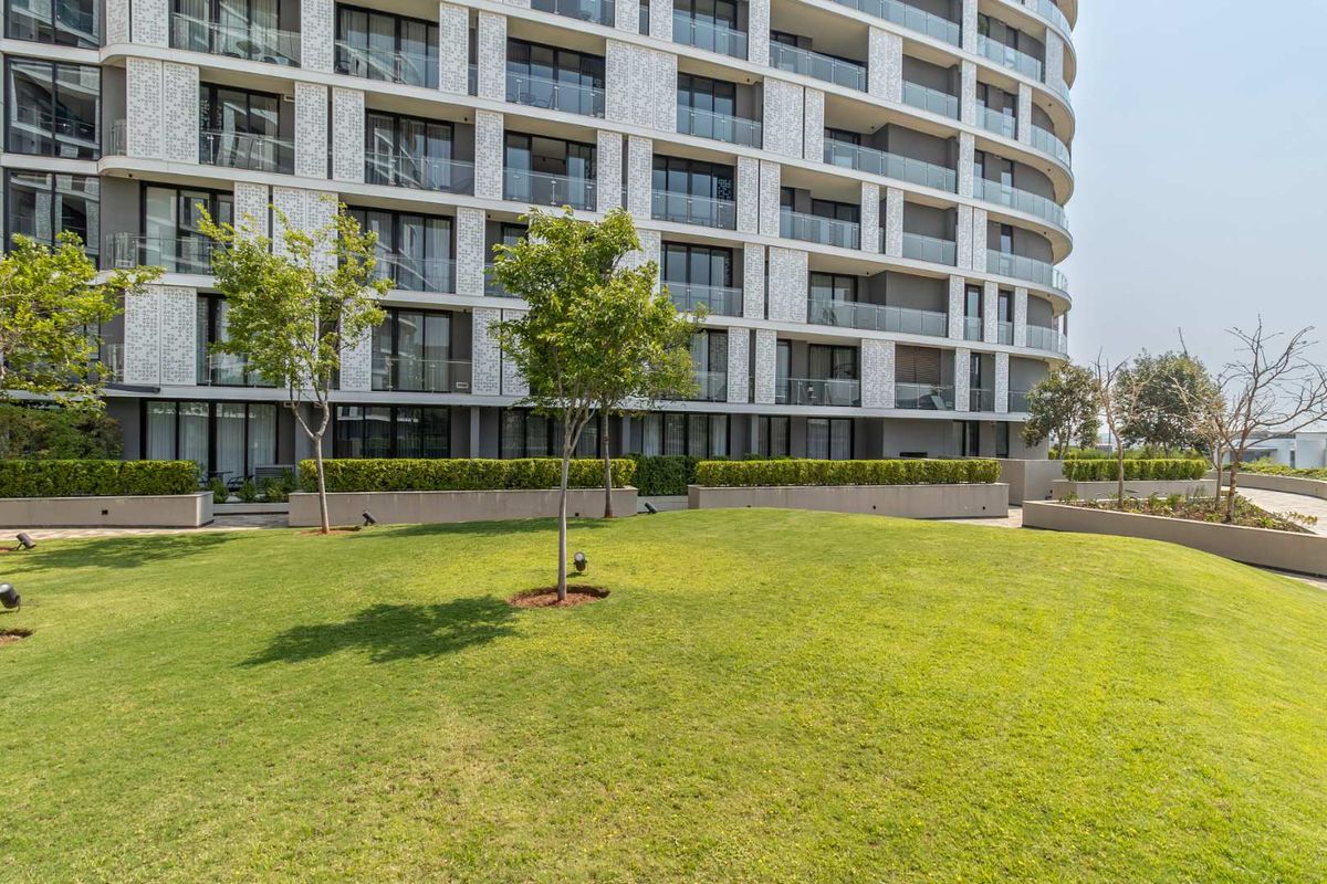A well-maintained landscaped garden area featuring a manicured lawn and retaining walls, set against the modern architectural facade of a multi-story apartment building.