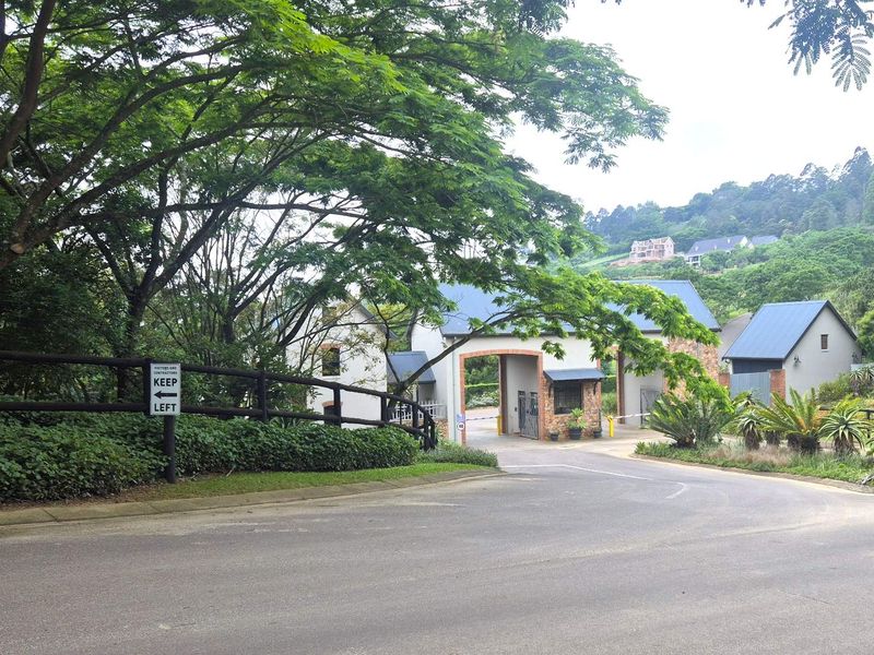 A view of the secure gated entrance to the residential estate, featuring a paved road, established trees, and a well-maintained security gatehouse.