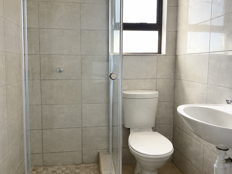 A clean, functional bathroom featuring a glass-enclosed shower, pedestal basin, and toilet with neutral wall and floor tiling.