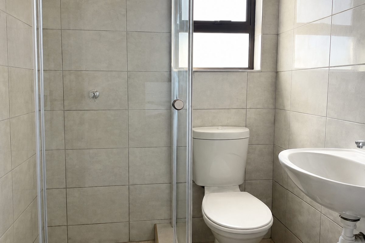 A clean, functional bathroom featuring a glass-enclosed shower, pedestal basin, and toilet with neutral wall and floor tiling.