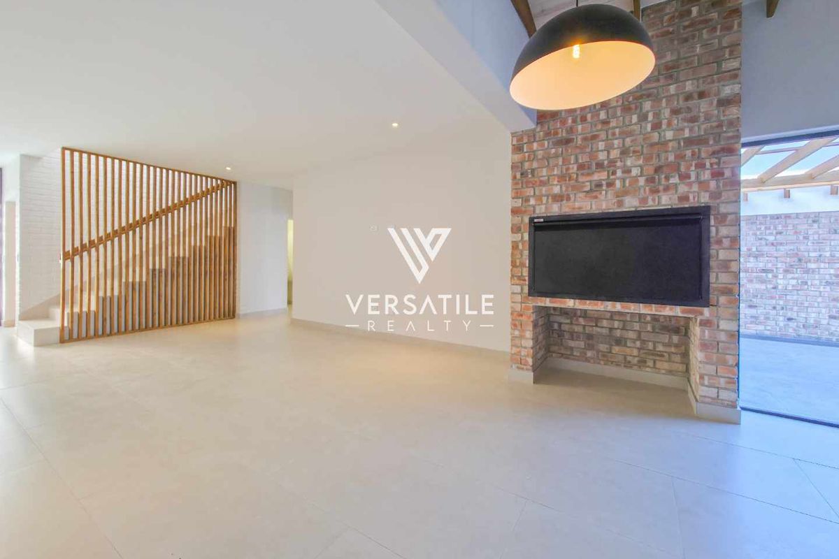 Spacious open-plan living area featuring a striking exposed brick fireplace, timber slat staircase partition, and modern tiled flooring with exposed ceiling beams.