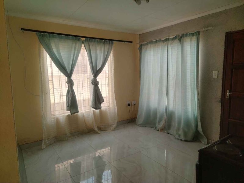 An empty living room featuring tiled flooring, security bars on windows, and a timber door, requiring cosmetic updates to the walls and finishes.