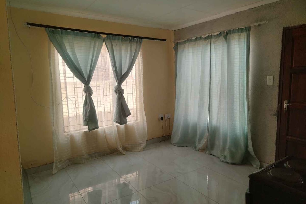 An empty living room featuring tiled flooring, security bars on windows, and a timber door, requiring cosmetic updates to the walls and finishes.