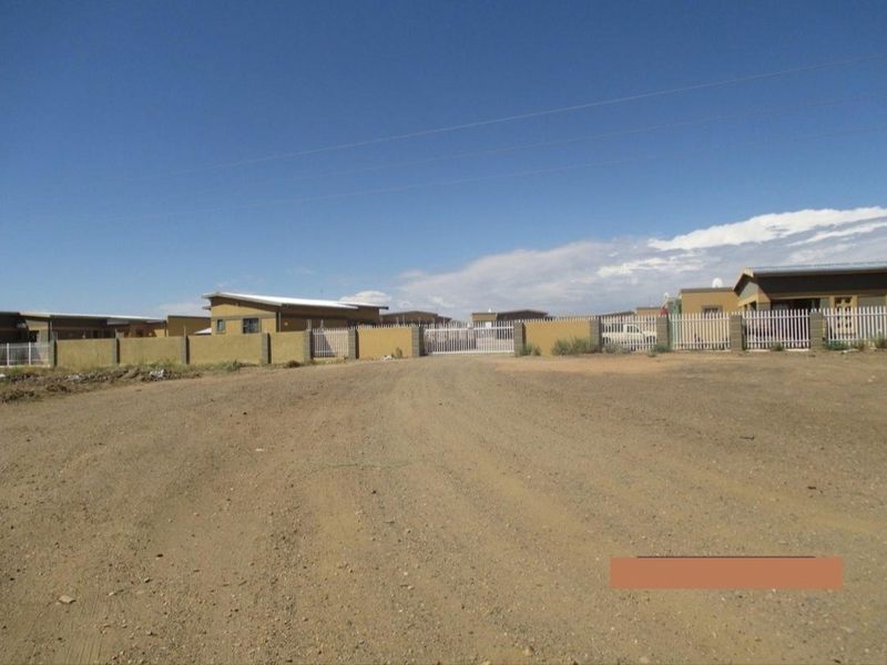 A street-level view of a residential development featuring perimeter fencing and a gated entrance with an unpaved access road.