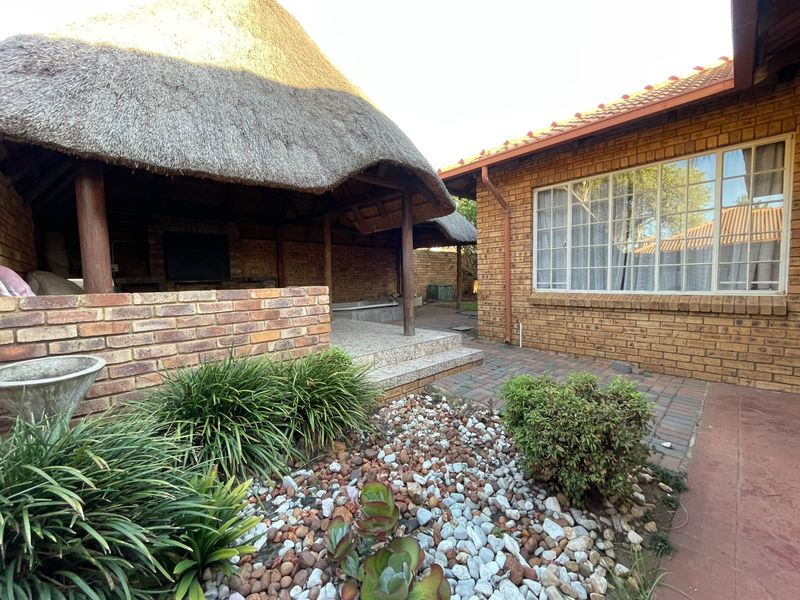 A covered outdoor patio area featuring a traditional thatch roof, built-in brick braai, and paved flooring, adjacent to the main house structure.