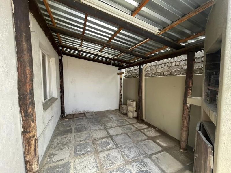 A covered outdoor patio area featuring a built-in braai, timber support beams, and paved flooring, offering a functional space for outdoor entertainment.