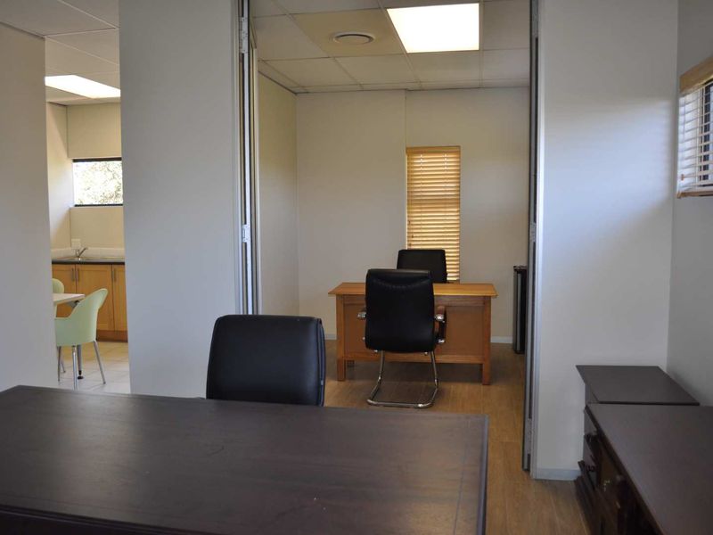 A functional office space featuring wood-look flooring, recessed lighting, and sliding partition doors, with an adjacent kitchenette area visible in the background.