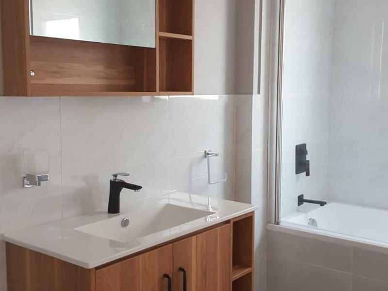 Modern bathroom featuring a wood-finish vanity with integrated sink, black fixtures, and a bathtub with a glass shower screen and rainfall shower head.