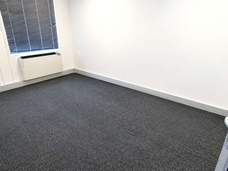A clean, empty office space featuring dark carpet flooring, a wall-mounted HVAC unit, and large windows with venetian blinds.