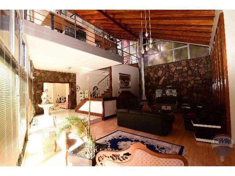 Spacious open-plan living area featuring a double-volume ceiling with exposed timber beams, stone feature walls, and a mezzanine level overlooking the main space.