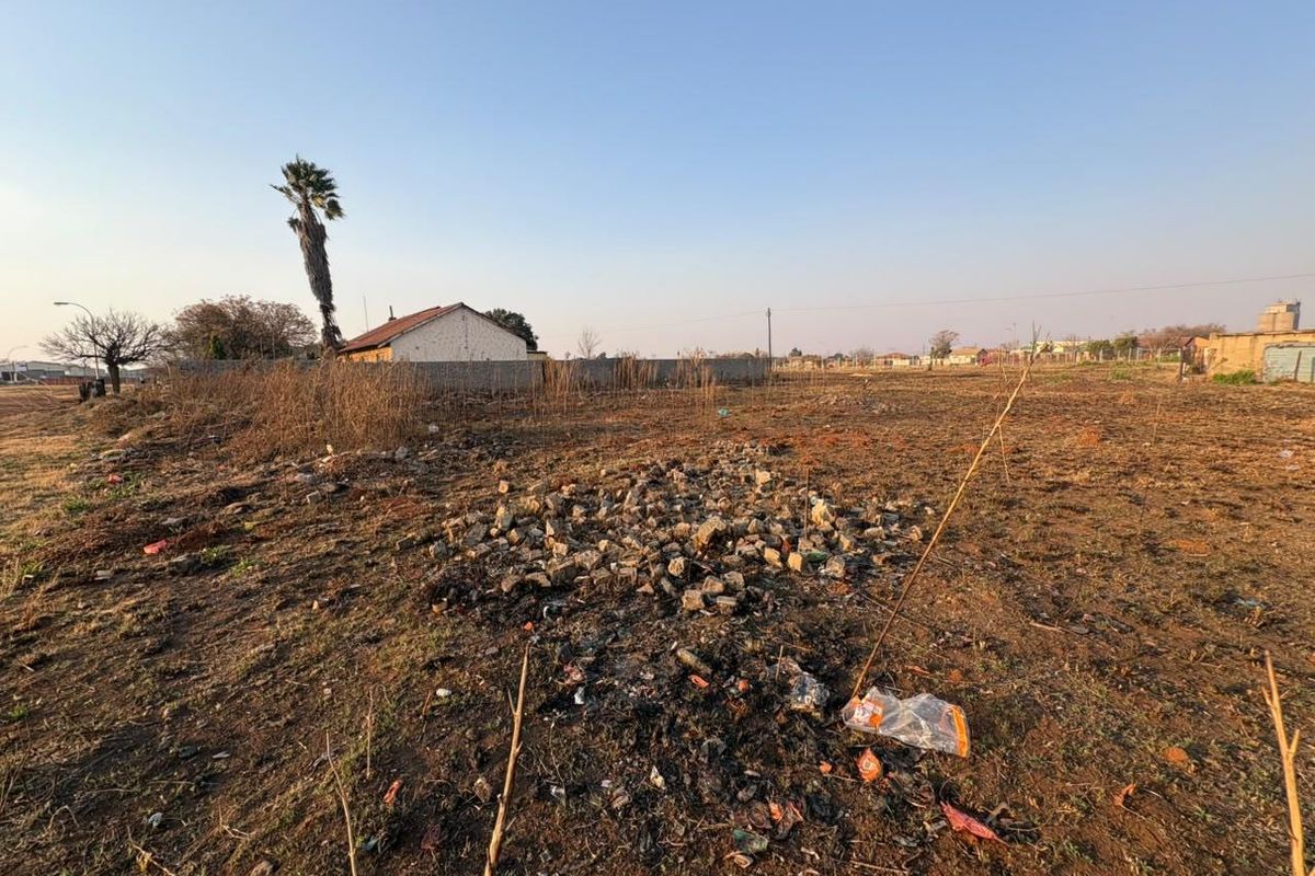 A vacant residential plot featuring dry vegetation and a boundary wall, offering a clear view of the surrounding neighborhood.