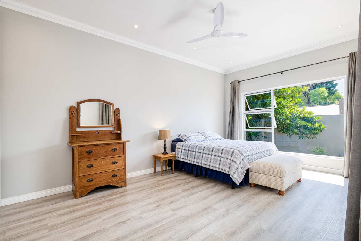 A bright and spacious bedroom featuring modern light-toned laminate flooring, recessed ceiling lighting, a white ceiling fan, and large windows overlooking the garden.