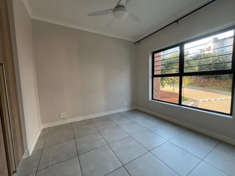 A bright, empty bedroom featuring neutral tiled flooring, a ceiling fan, and a large window with security bars.