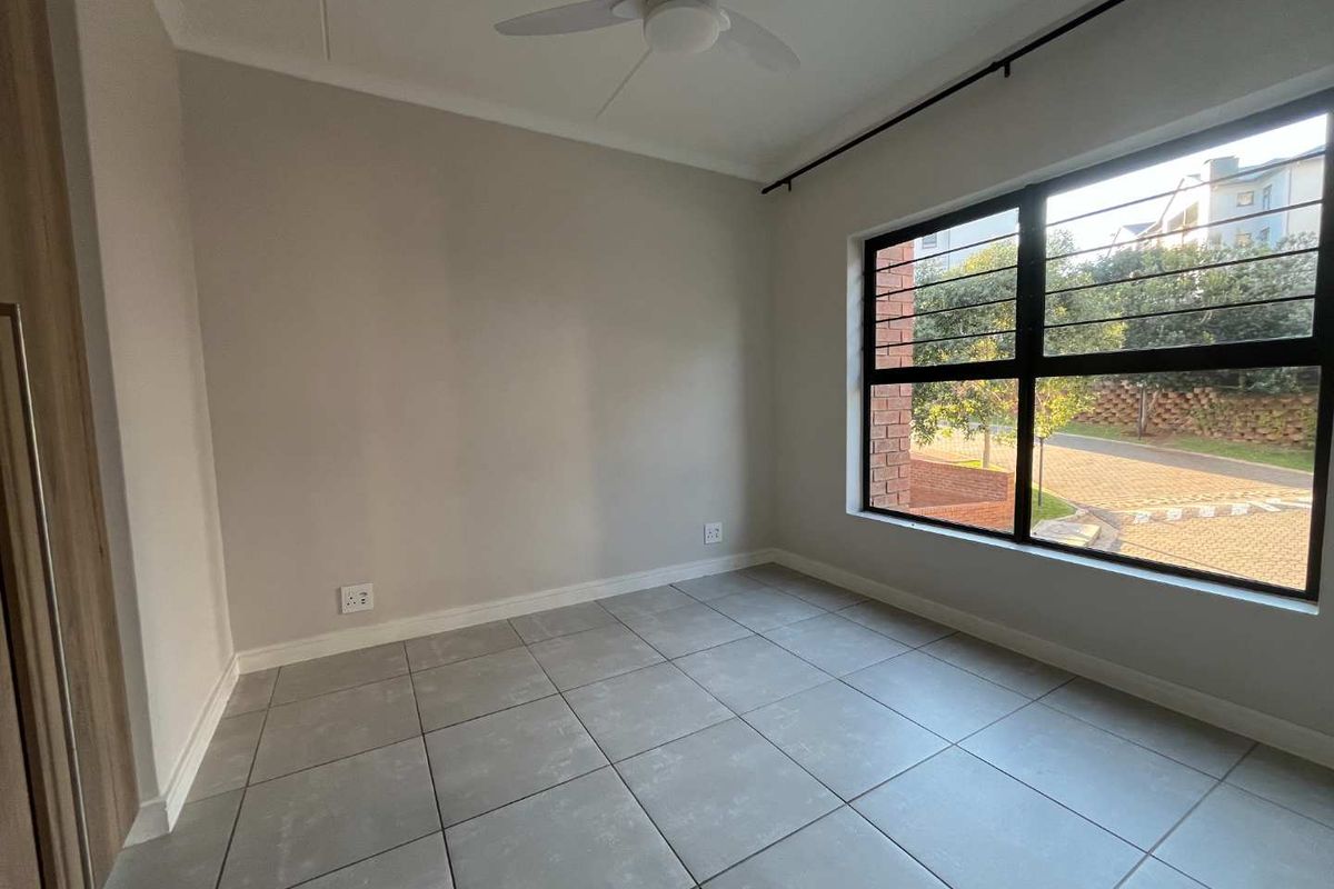 A bright, empty bedroom featuring neutral tiled flooring, a ceiling fan, and a large window with security bars.