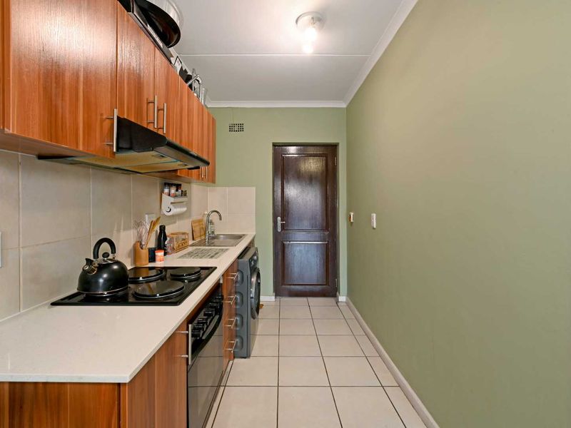 A functional galley-style kitchen featuring wood-finish cabinetry, white countertops, tiled splashbacks, and integrated cooking appliances with tiled flooring.