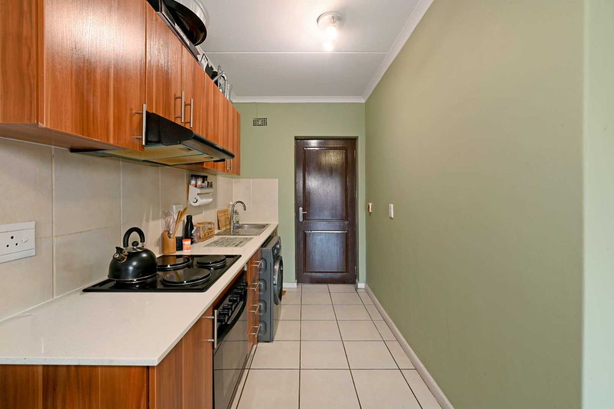 A functional galley-style kitchen featuring wood-finish cabinetry, white countertops, tiled splashbacks, and integrated cooking appliances with tiled flooring.