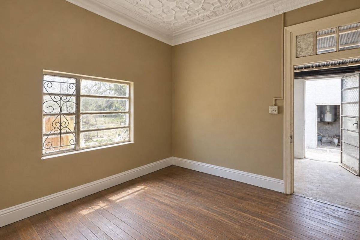 An empty bedroom featuring original wooden flooring, an ornate ceiling, and a steel-framed window with security bars, leading to an adjacent utility area.