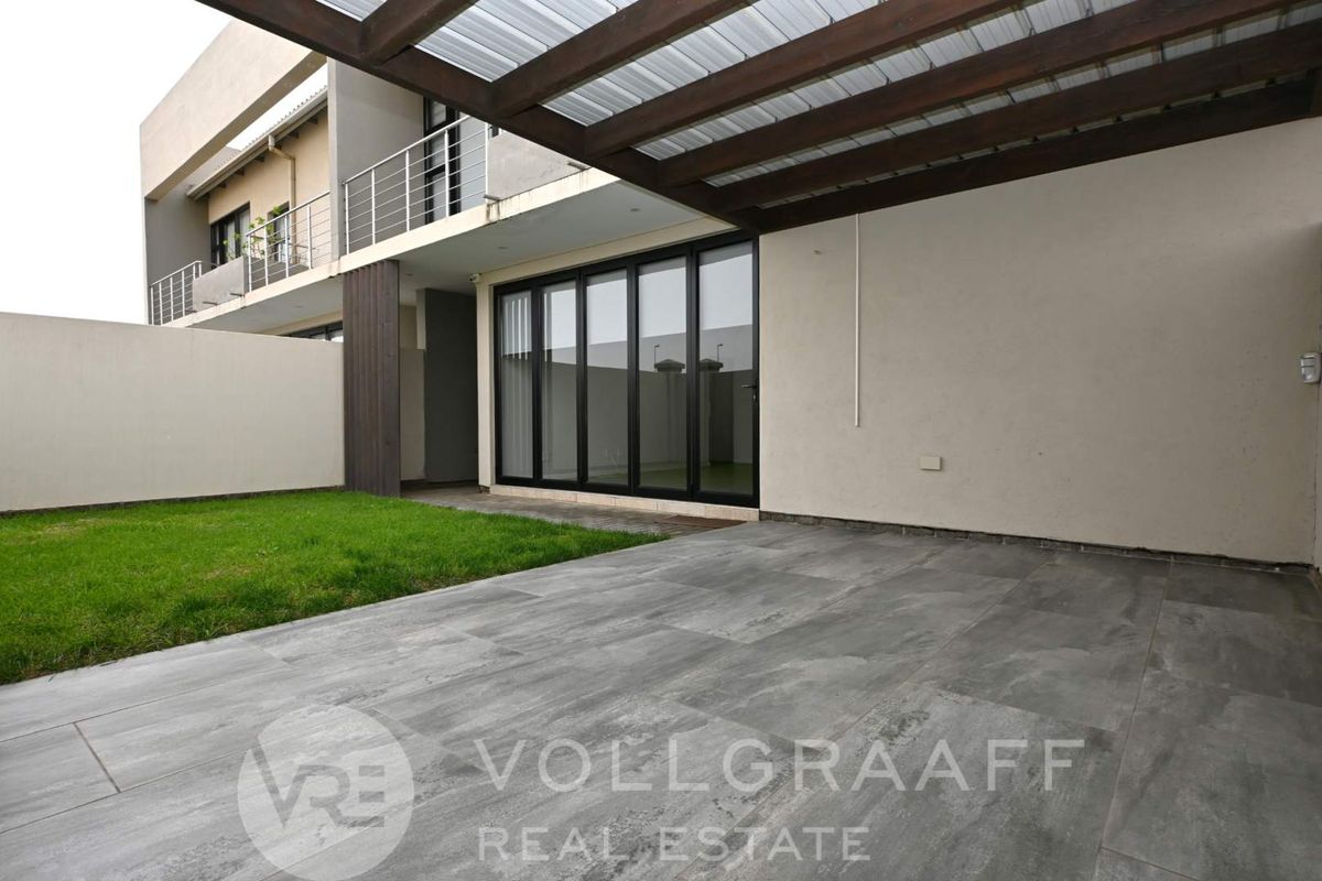 A modern rear exterior view featuring a covered patio with tiled flooring, a small lawn, and large black-framed sliding glass doors leading indoors.