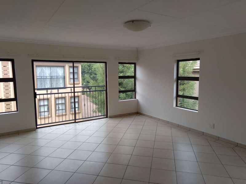 A bright, empty living room featuring neutral tiled flooring, large windows, and a sliding glass door leading to a balcony.