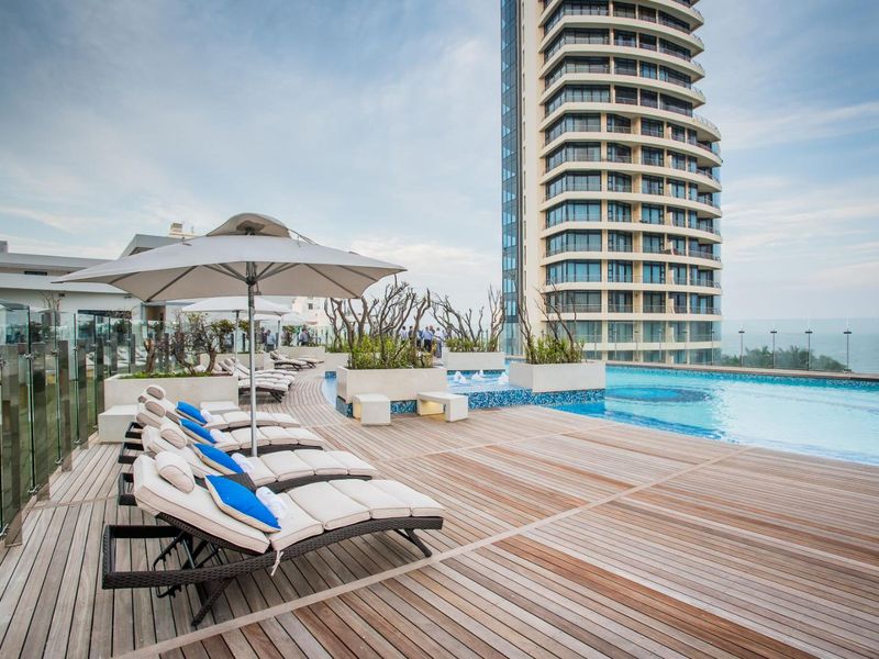 A luxury rooftop swimming pool area featuring timber decking, glass balustrades, and built-in planters with views of the high-rise building and ocean.
