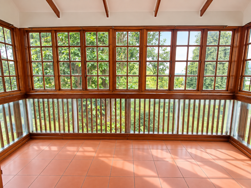 A bright, enclosed sunroom or patio featuring terracotta floor tiles, exposed timber rafters, and extensive timber-framed windows overlooking the garden.